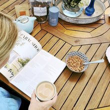 Load image into Gallery viewer, A view from above of a woman reading a magazine with a cup of coffee. A bowl of muesli sits on the table to her right.
